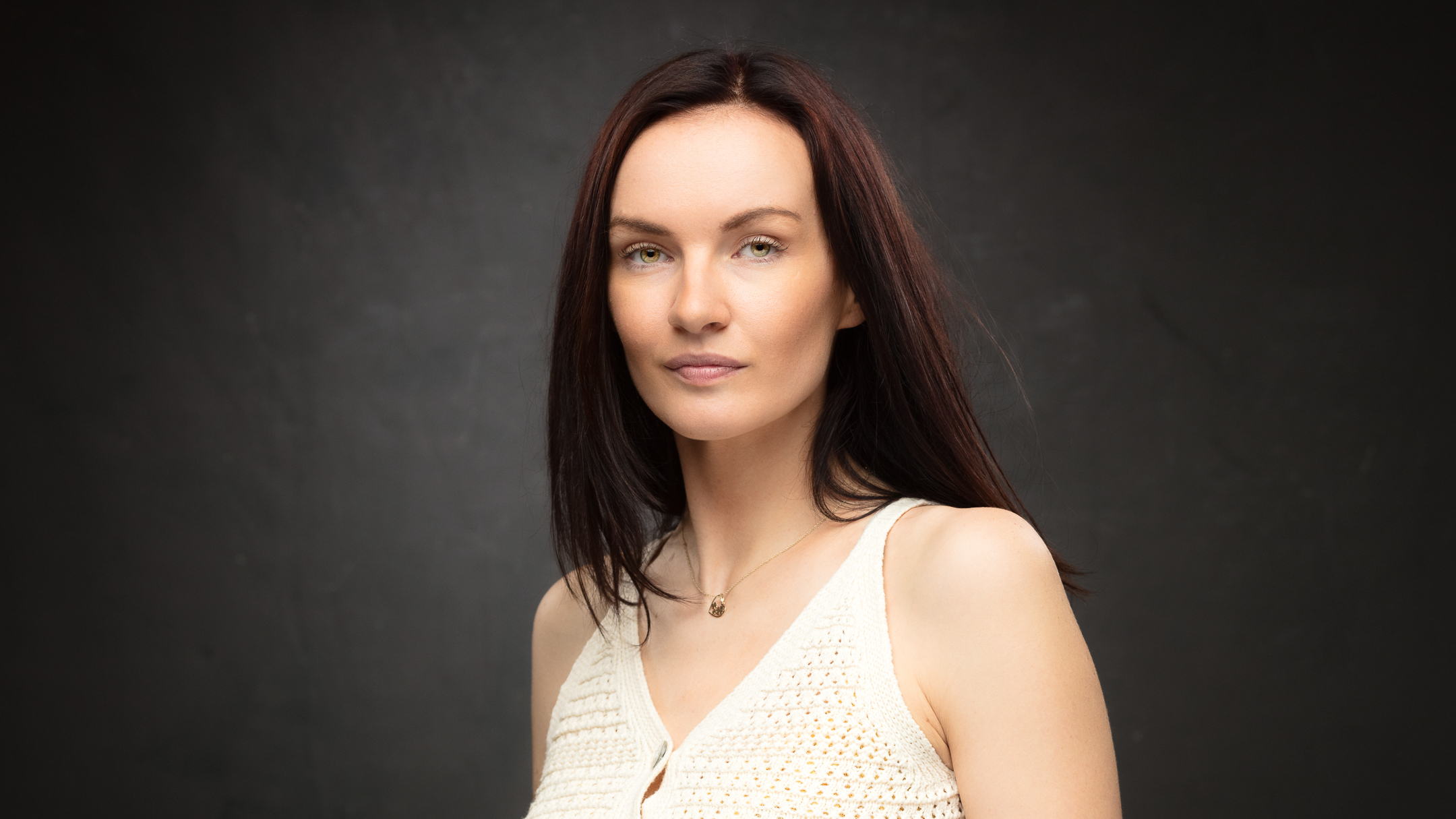 Studio portrait of a woman with long dark hair in a cream top, against a dark grey backdrop