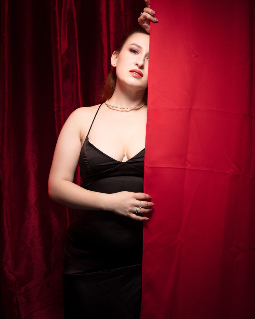 Woman in black dress peeks from behind red curtain.