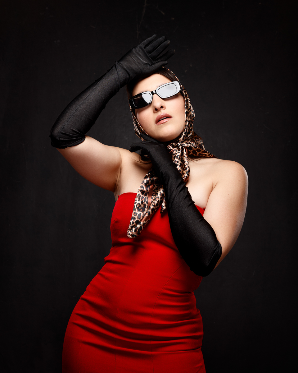Retro pin-up style studio portrait of Mariana in a red dress with a leopard-print headscarf, sunglasses and black gloves