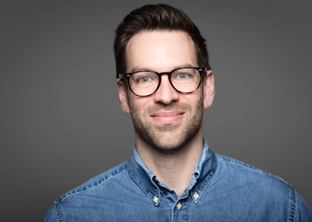 Man in glasses and denim shirt smiling.