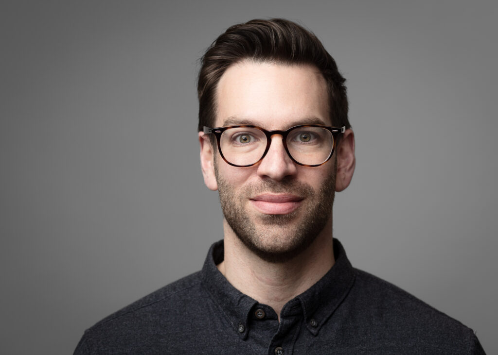 Smiling man with glasses and dark shirt.