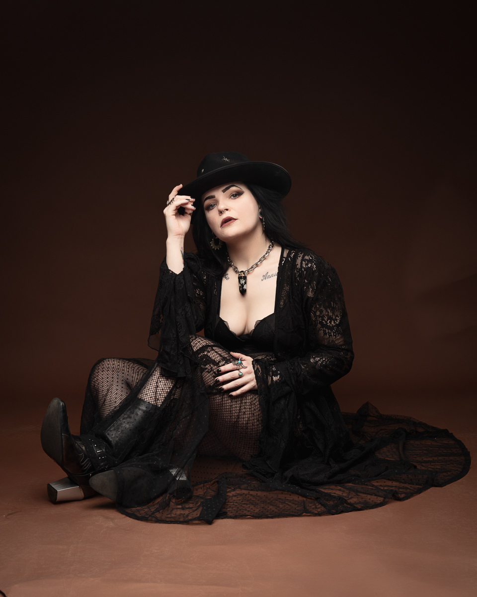 Woman in black lace outfit, seated and wearing hat.