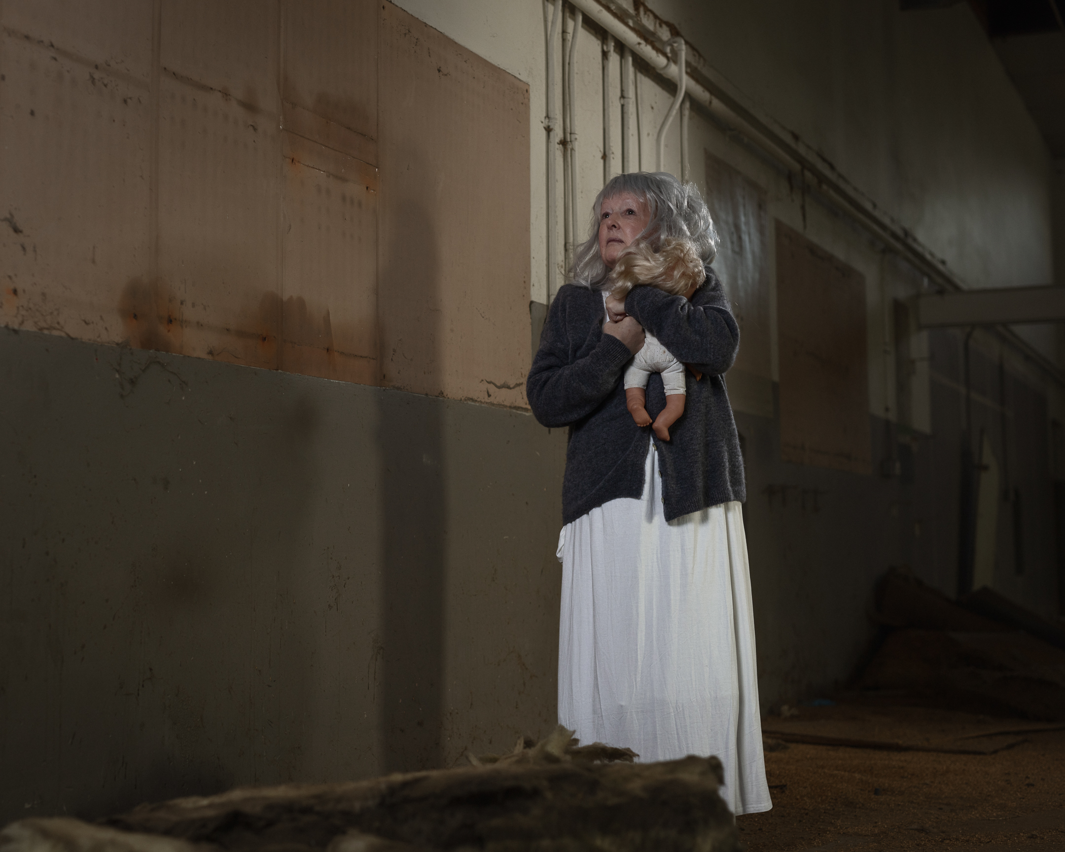Elderly woman holding doll in dim corridor.
