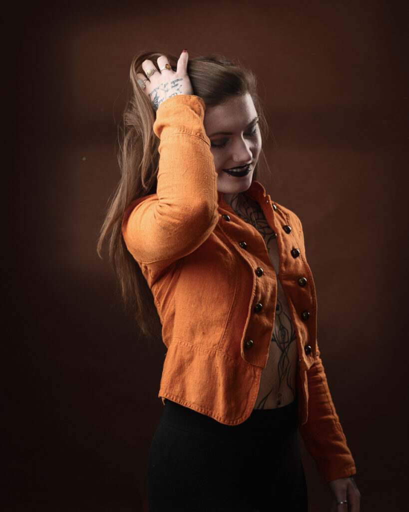 Person with tattoos wearing orange jacket posing confidently.