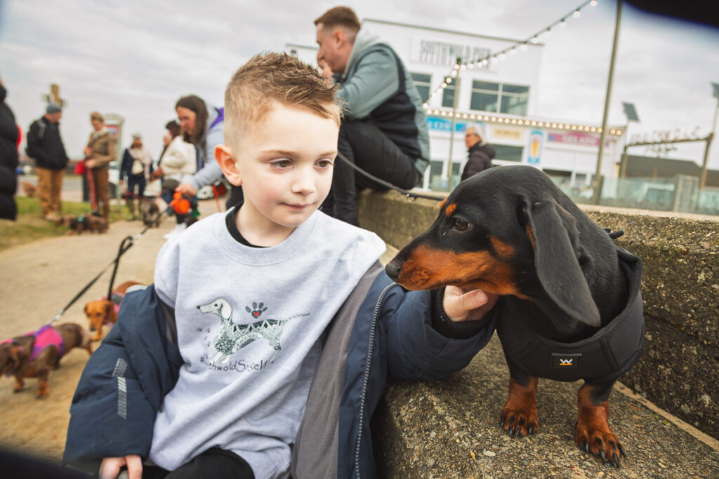 Child petting a dachshund on a seaside walkway.