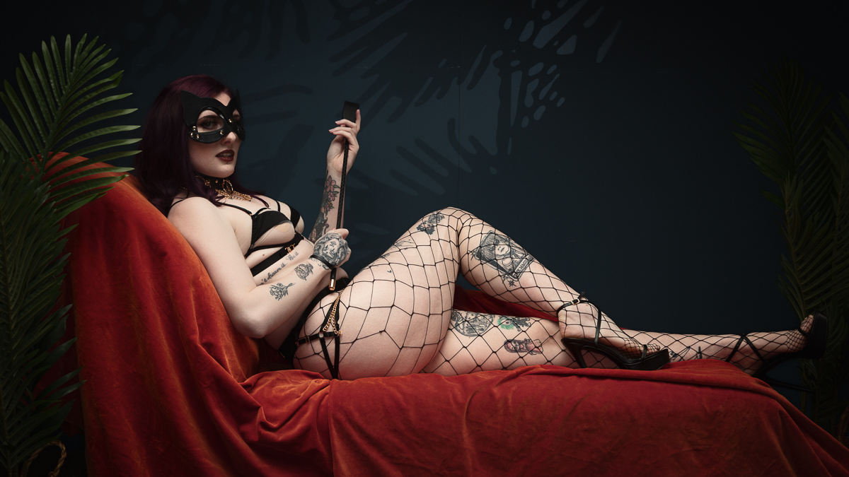 Tattooed woman in lingerie and mask reclines on sofa.