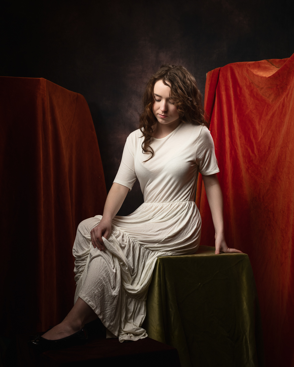 Woman in white dress, seated, against red drapery.