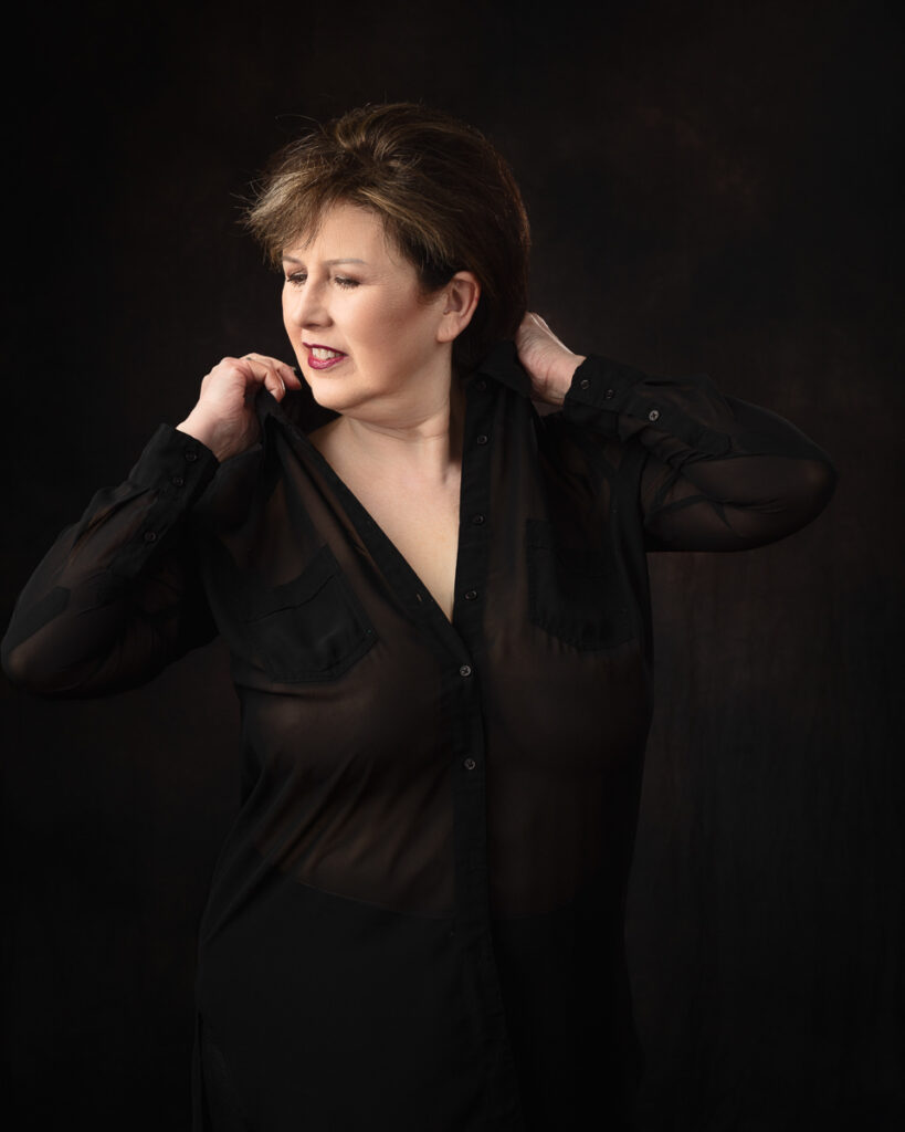 Person posing in black sheer shirt, dark background.