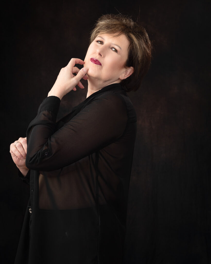 Person posing stylishly in black outfit against dark backdrop.