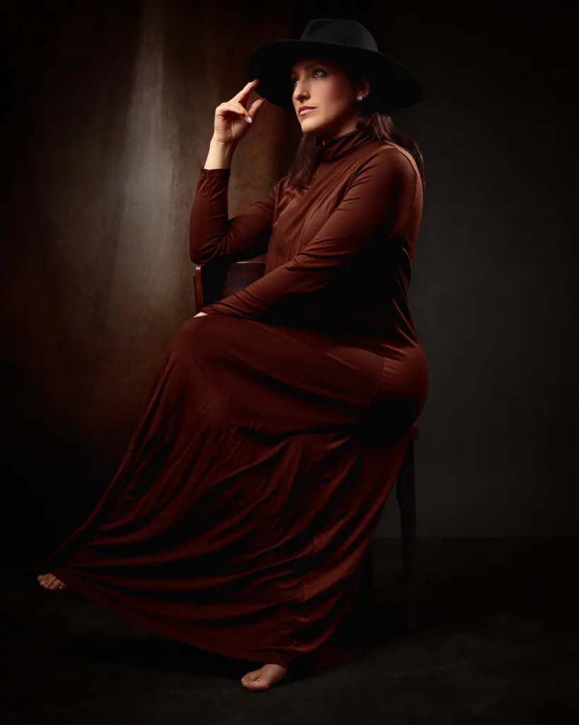Elegant woman in dark hat and dress seated.