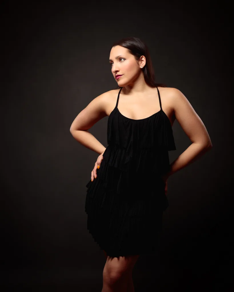 Woman in black dress posing against dark background.