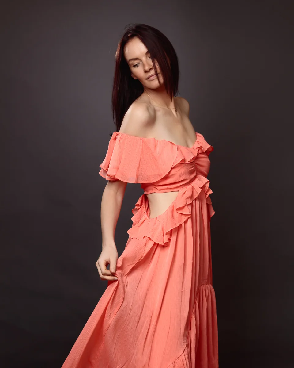 Woman in coral off-shoulder ruffled dress, elegant pose.