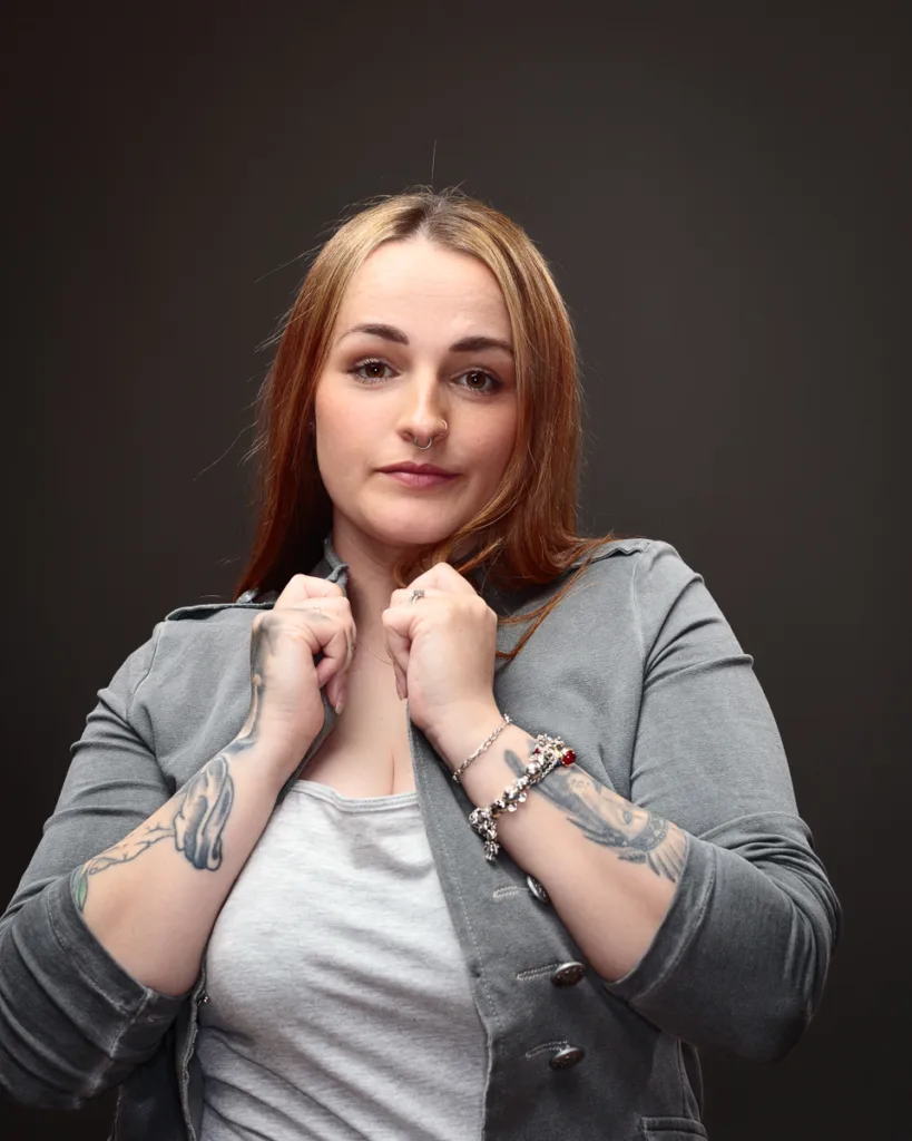 Woman in grey jacket with tattoos poses confidently.