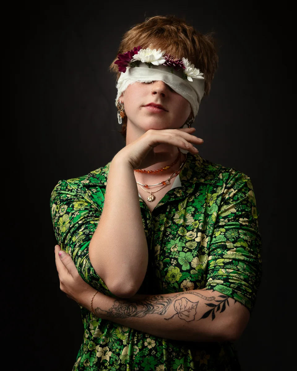 Person with floral blindfold, arm tattoos, green dress