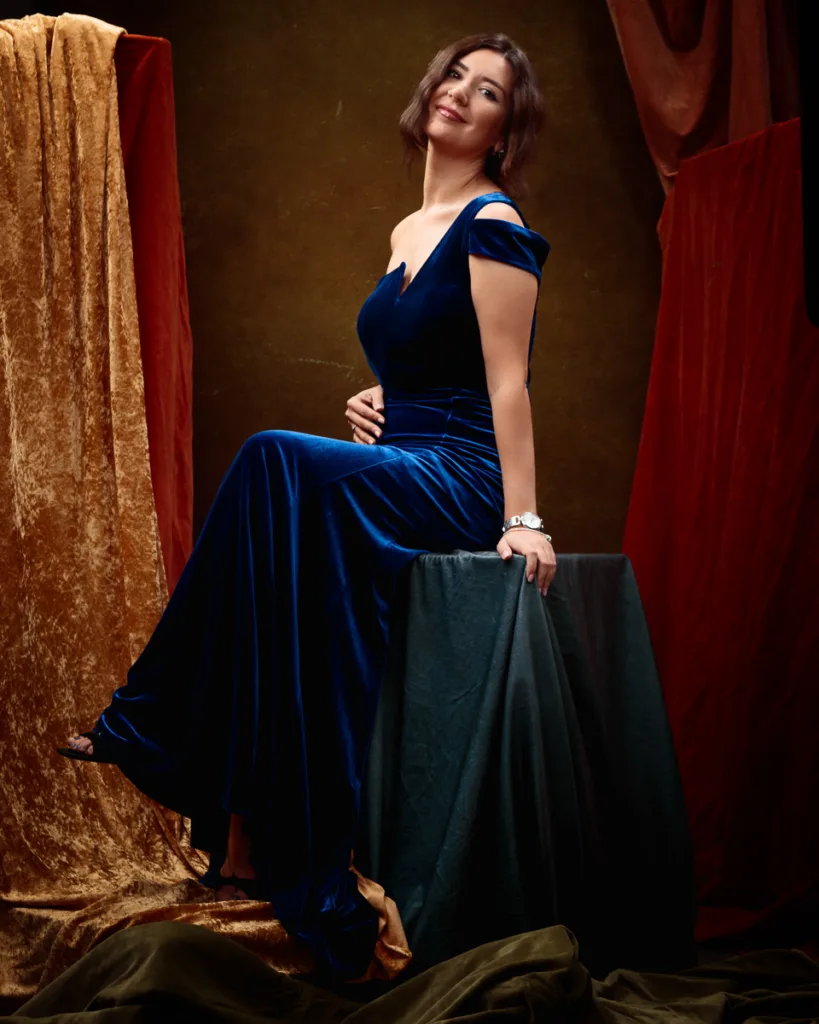 Woman in blue velvet dress seated against draped fabric.