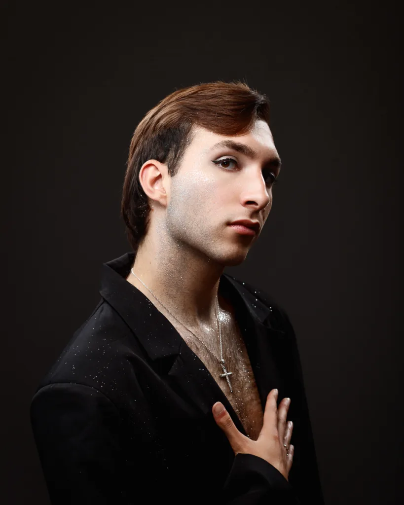 Person with makeup, wearing black blazer, hand on chest.