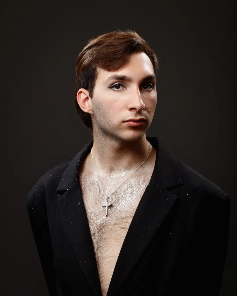 Person in glitter with cross necklace, wearing black blazer.