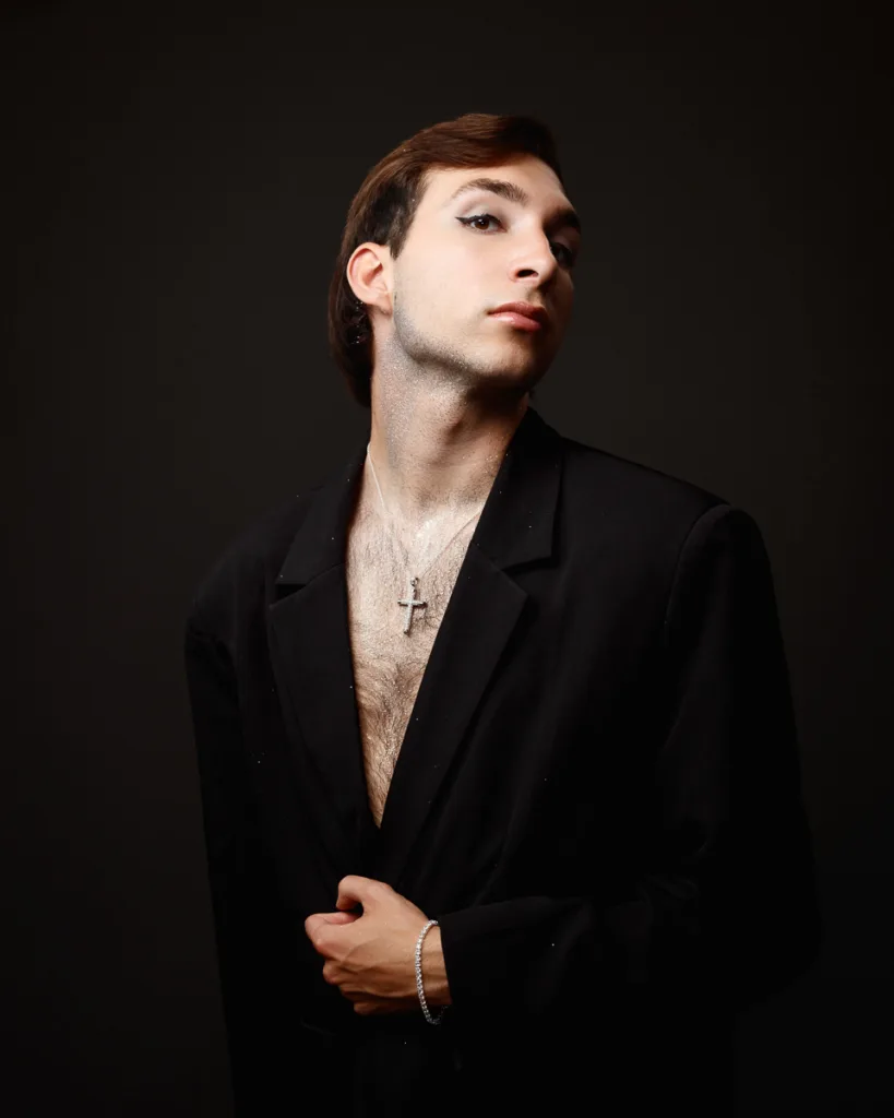 Person posing confidently in black suit, dark background.
