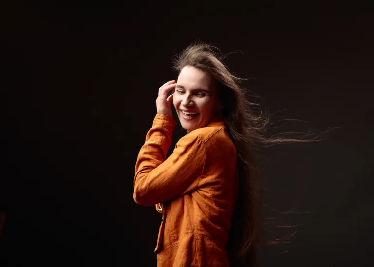 Woman smiling with windblown hair in orange jacket.