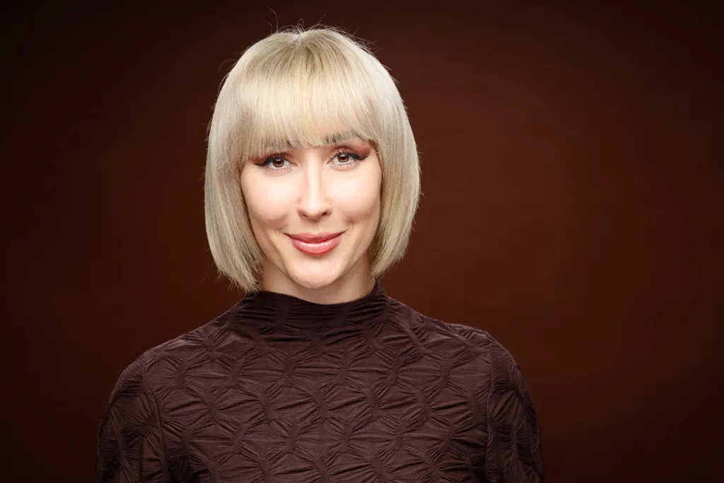 Smiling person with short blonde hair, brown background.