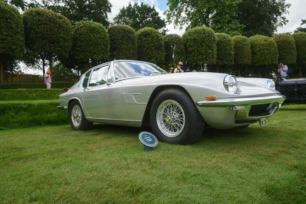 Classic silver car on grass field with people.
