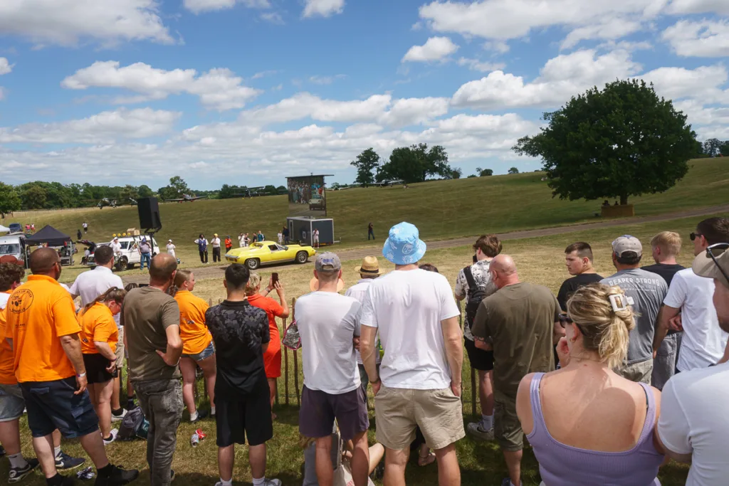 Outdoor event with crowd and yellow car, sunny day.