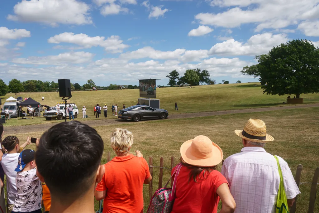Crowd watches car at outdoor event on grassy field.