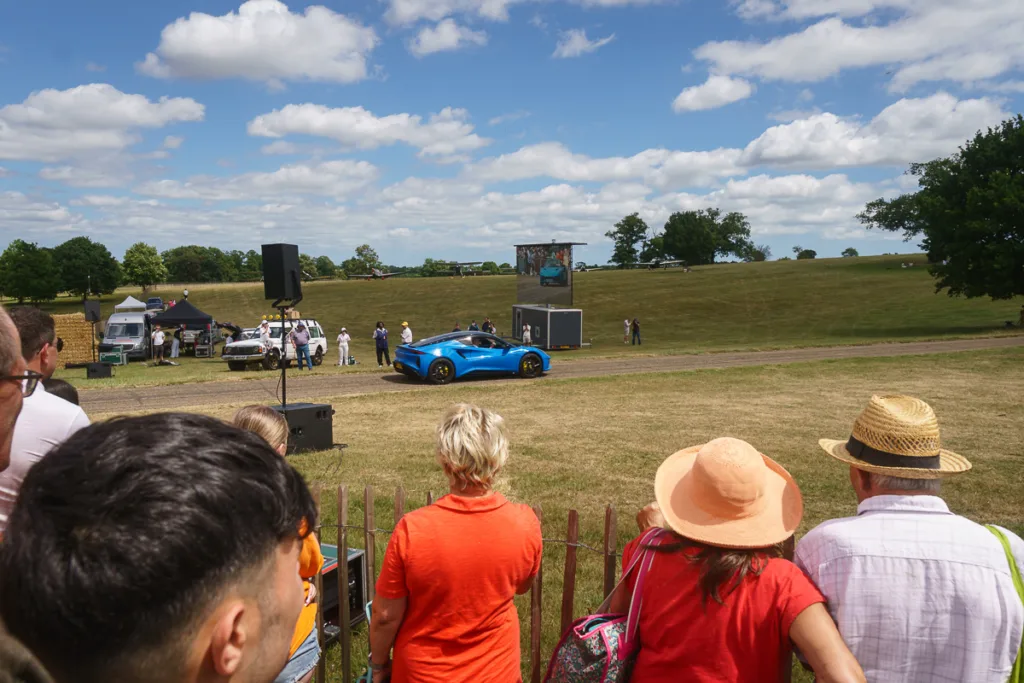 Crowd watches blue sports car on grassy field.