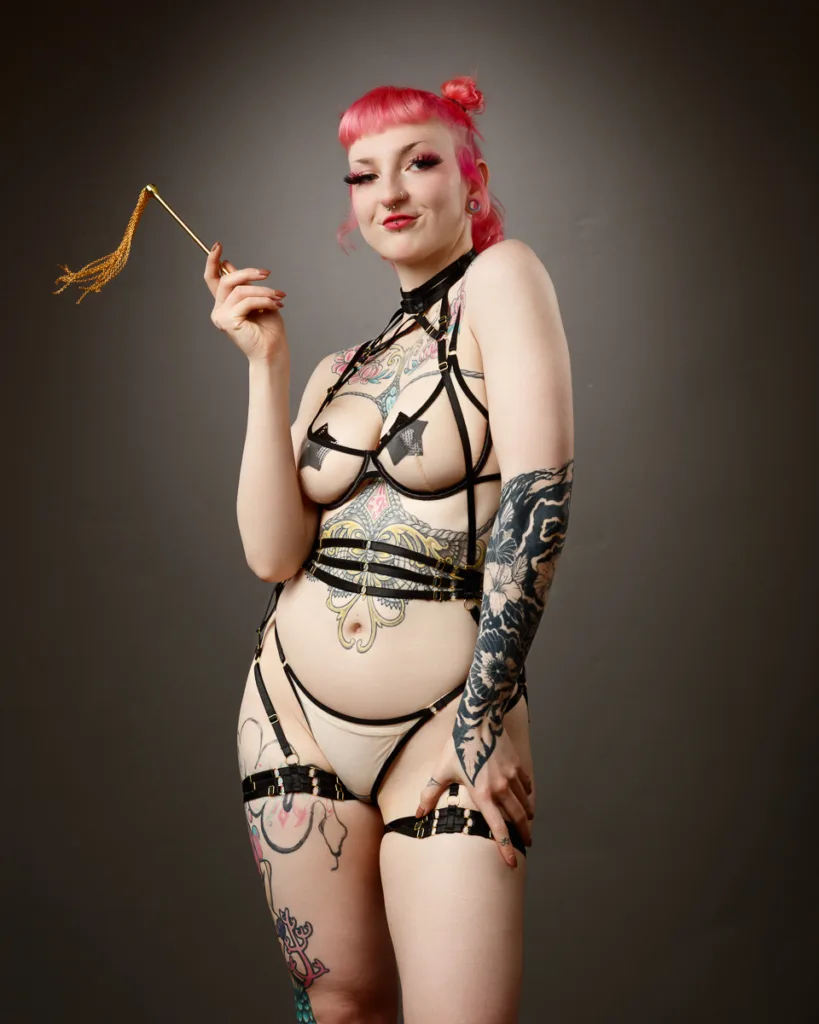 Kinki Couture studio shoot, pink-haired tattooed model wearing black harness lingerie and holding a gold tassel