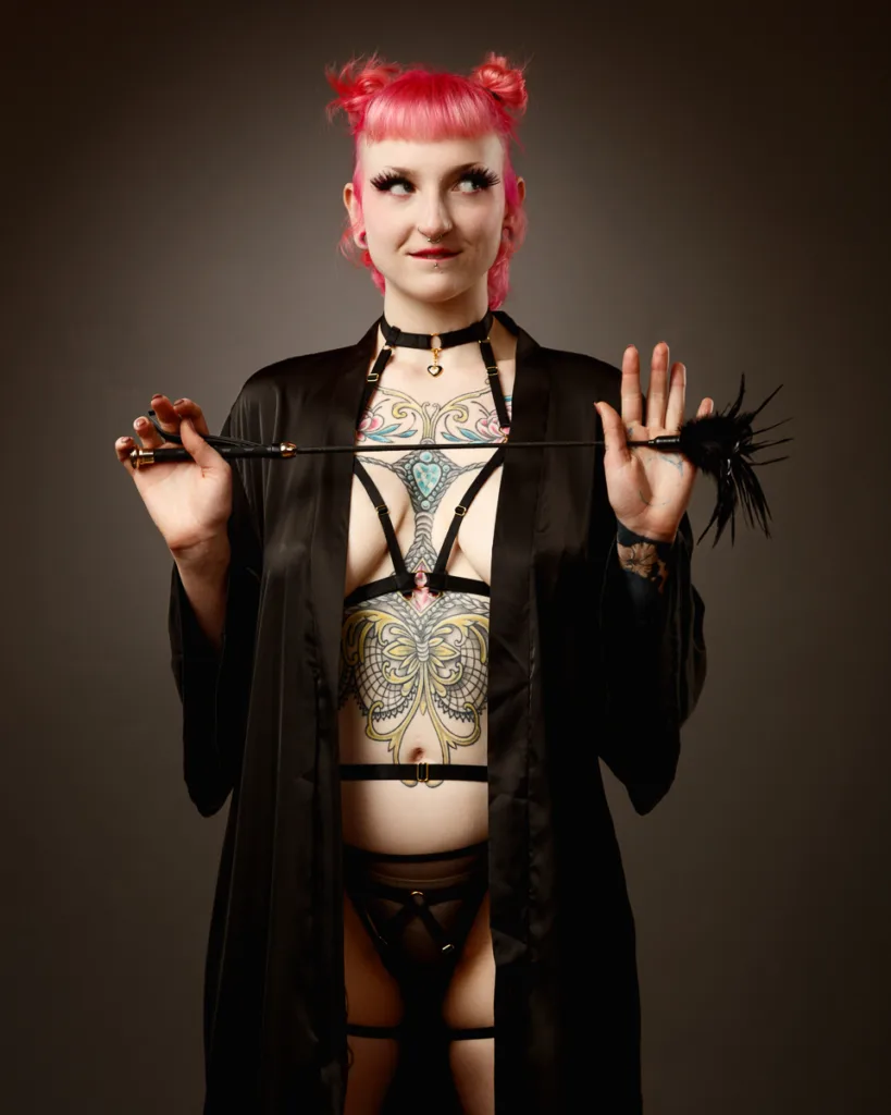 Person with tattoos, black robe, and pink hair holding whip.