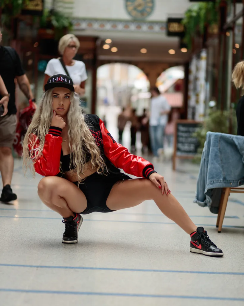 Woman squatting in arcade, wearing red jacket, black hat.