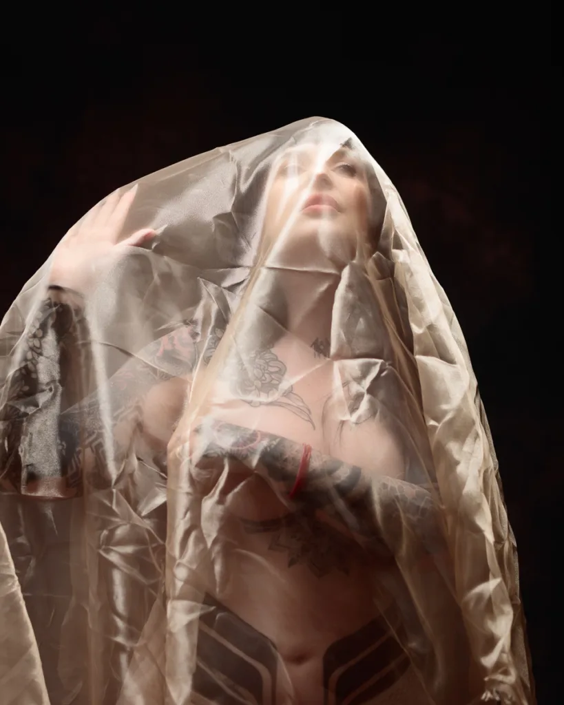 Tattooed person under sheer fabric against dark background.