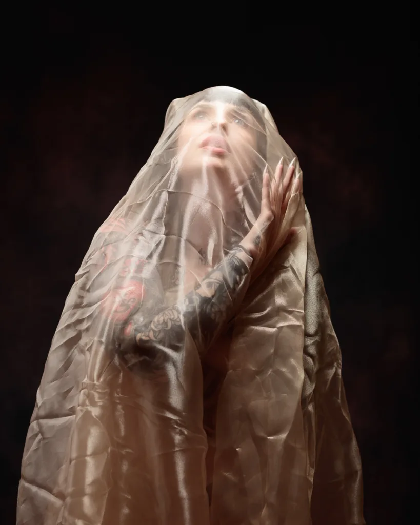 Tattooed person draped in sheer fabric, gazing upwards.