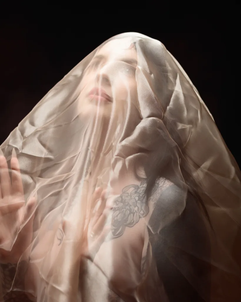 Person veiled in sheer fabric with tattoos visible.