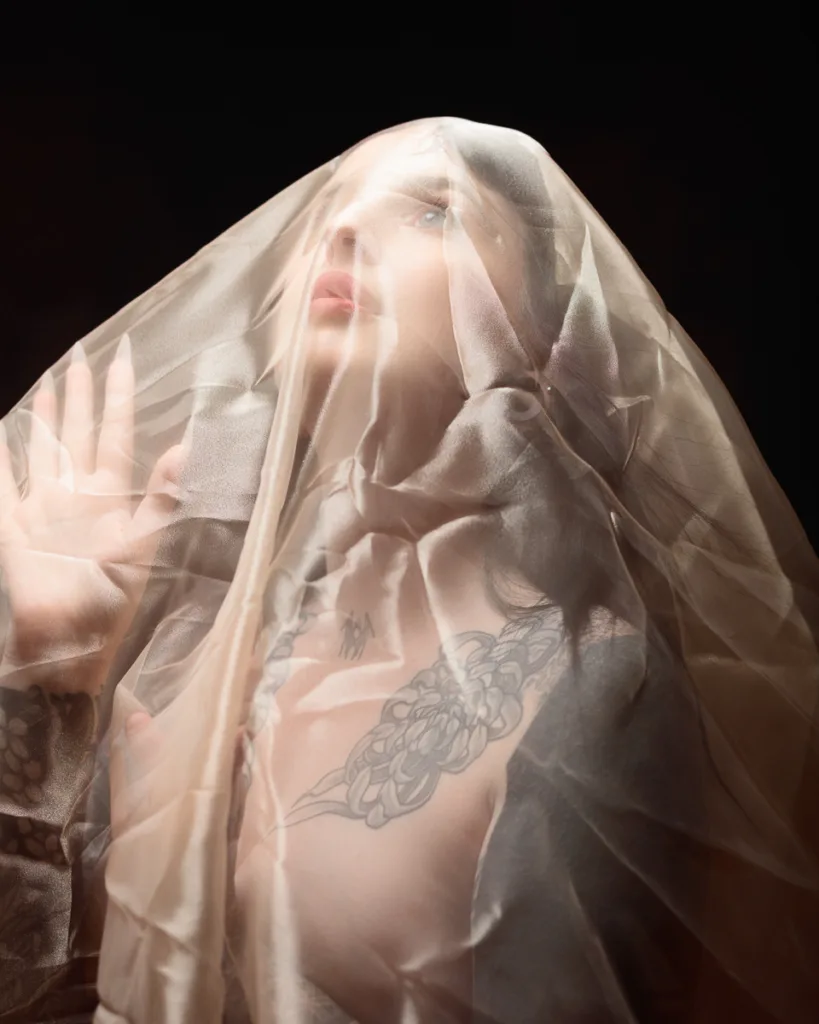Woman under sheer fabric with tattoos, gazing upward.