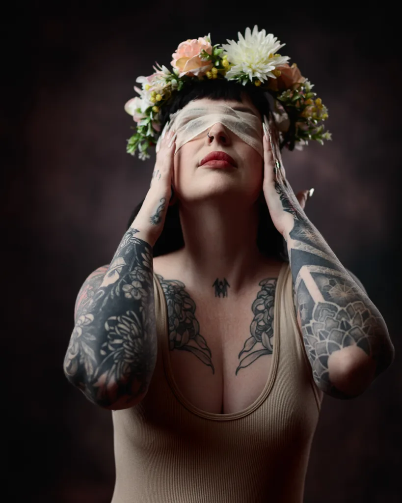 Tattooed person wearing floral crown and blindfold.