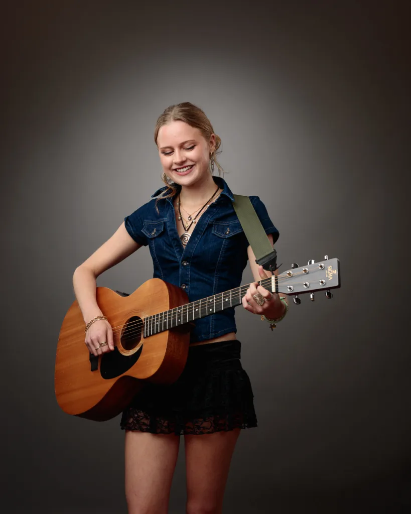 Woman playing guitar, smiling, in denim shirt.