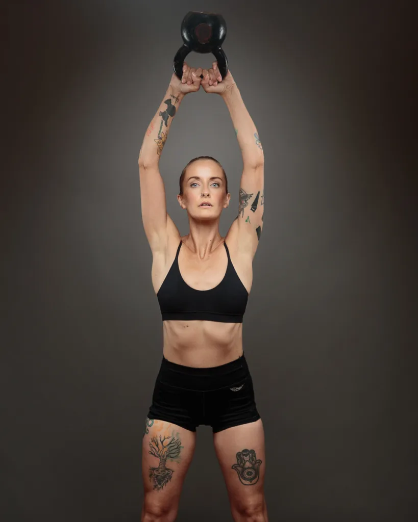 Woman lifting kettlebell, displaying tattoos and strength.