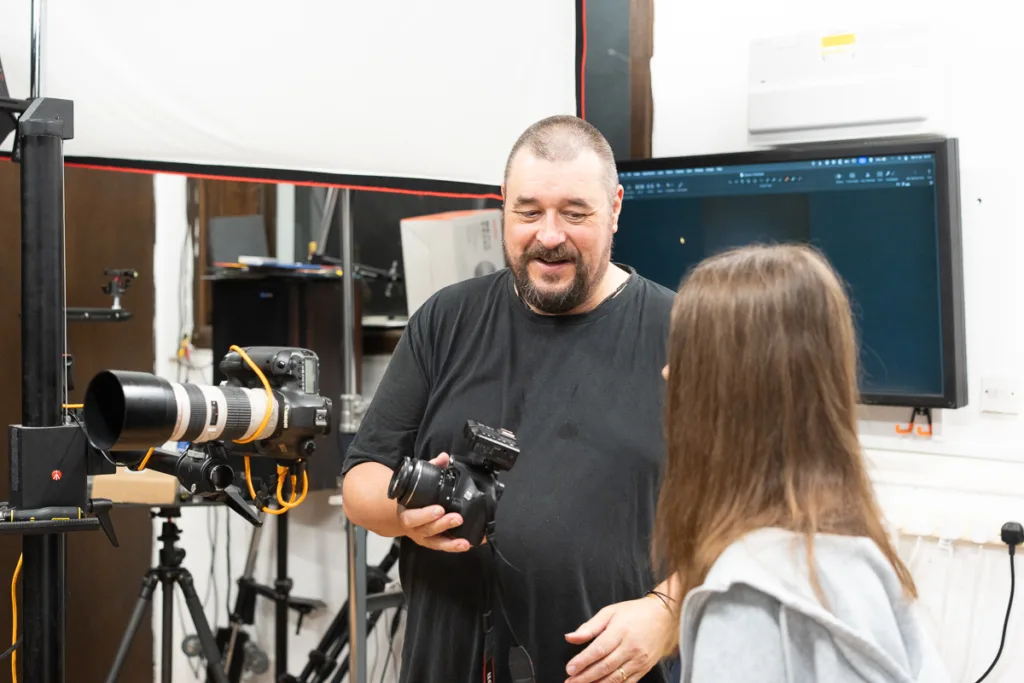 Photographer instructs student in studio with camera equipment.
