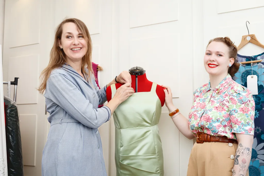 Two women adjusting dress on mannequin, smiling.