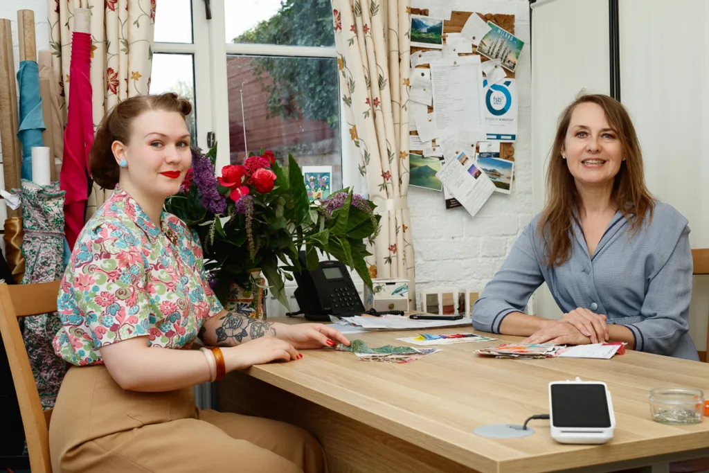 Two women sitting at a table with fabrics.