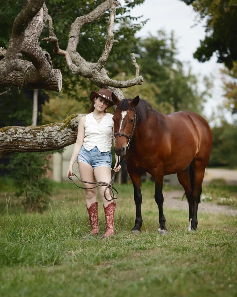 Person in cowboy attire with horse under tree.