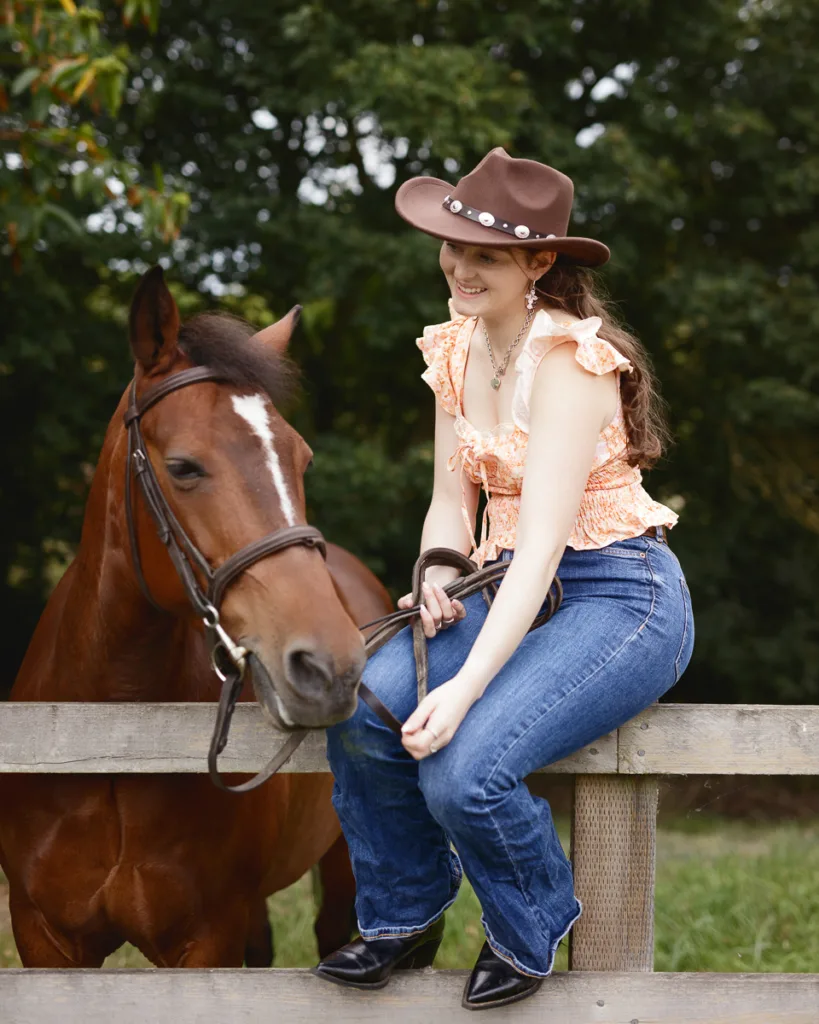 Woman in cowboy hat sitting on fence with horse.