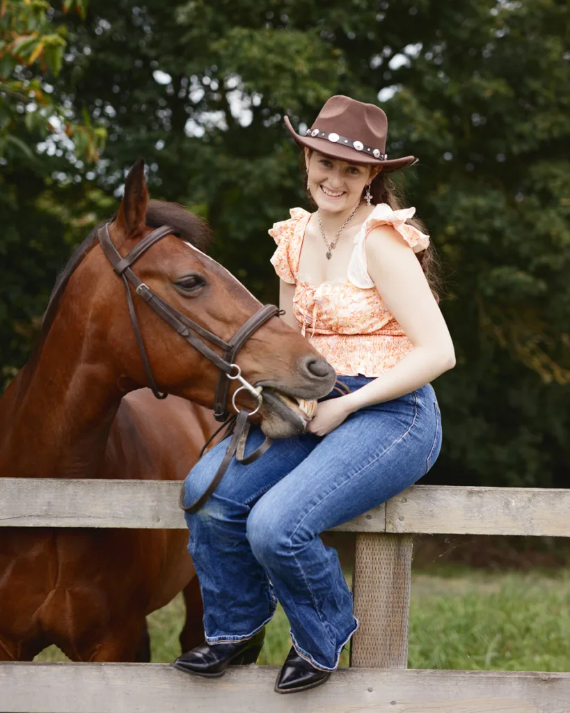 Smiling cowgirl sitting on fence with horse.