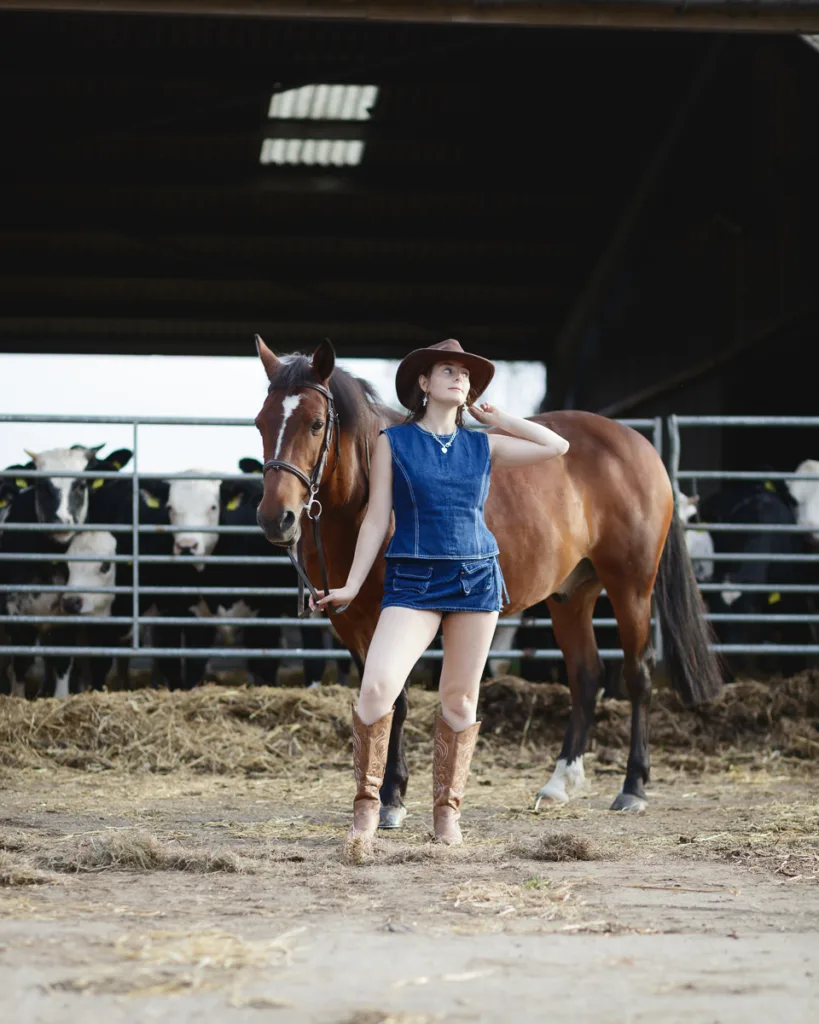 Woman with horse in farmyard, cows in background.