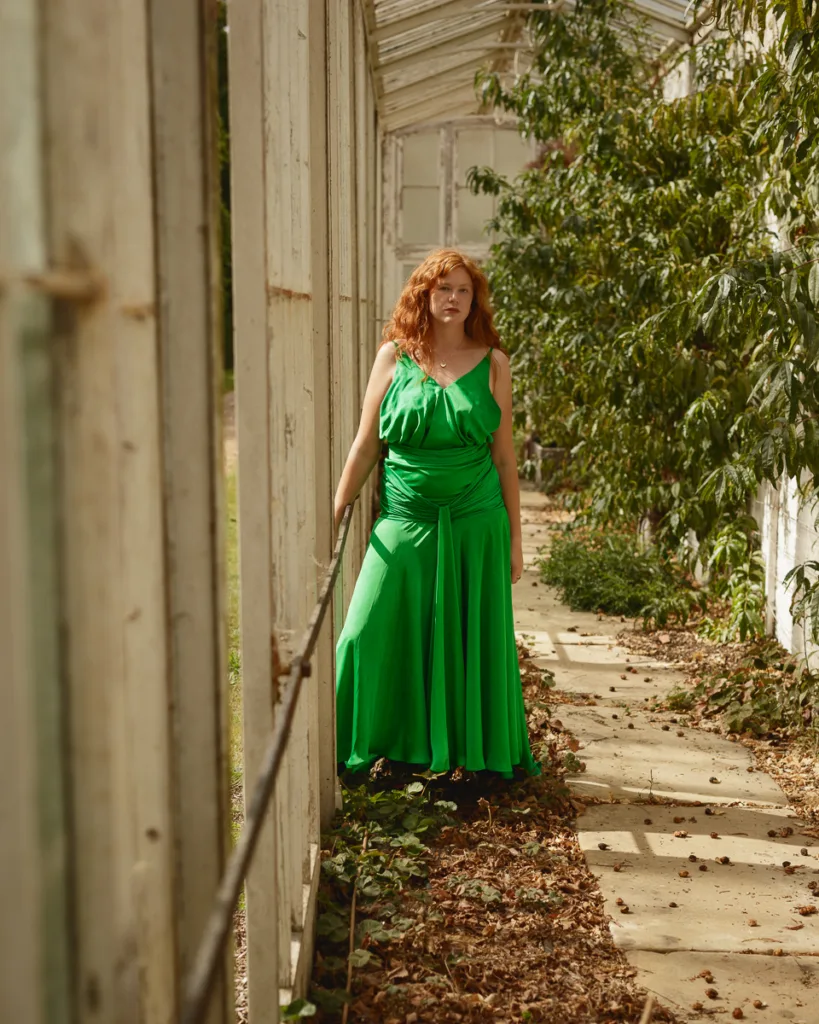 Woman in green dress walking in leafy corridor.