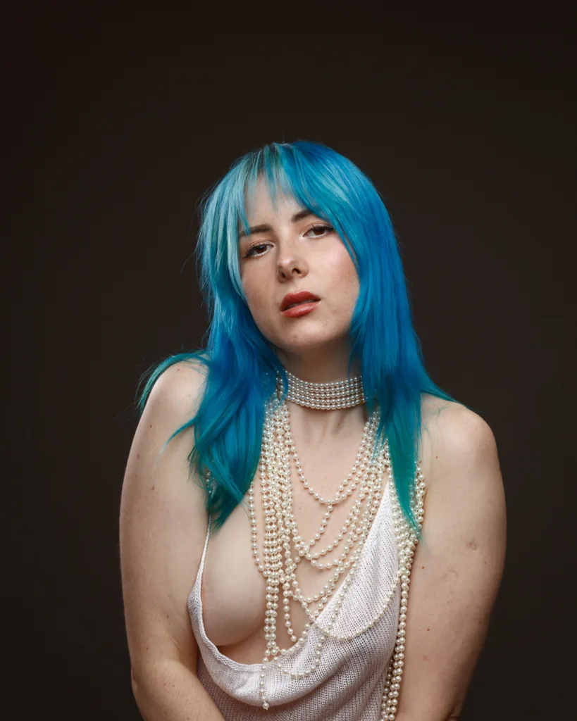 Person with blue hair wearing pearls and white attire.