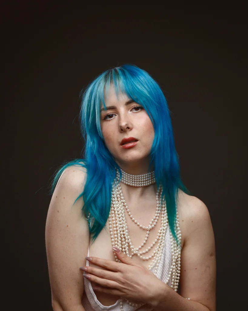 Person with blue hair, pearl necklaces, dark background.