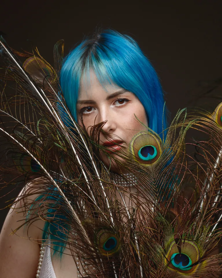 Person with blue hair behind peacock feathers.