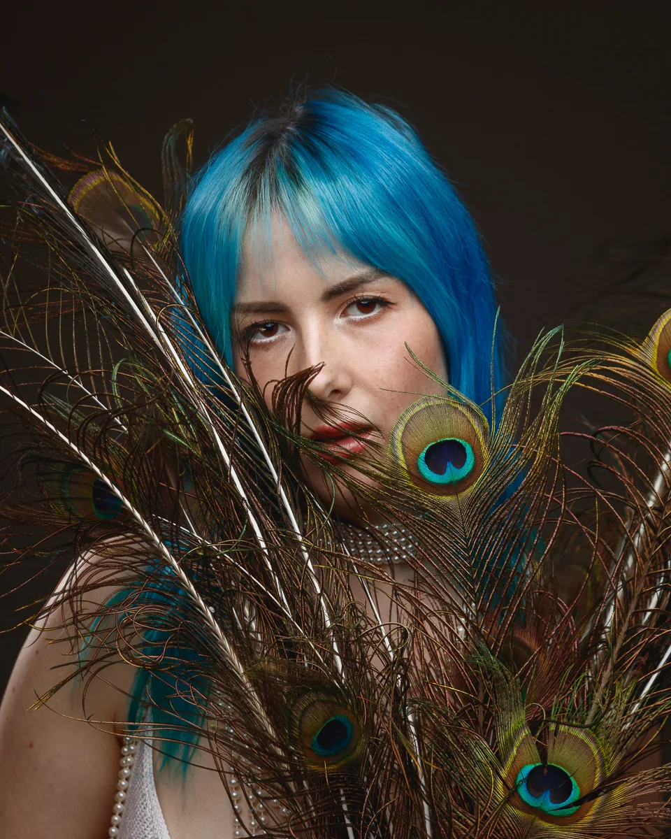Person with blue hair behind peacock feathers.